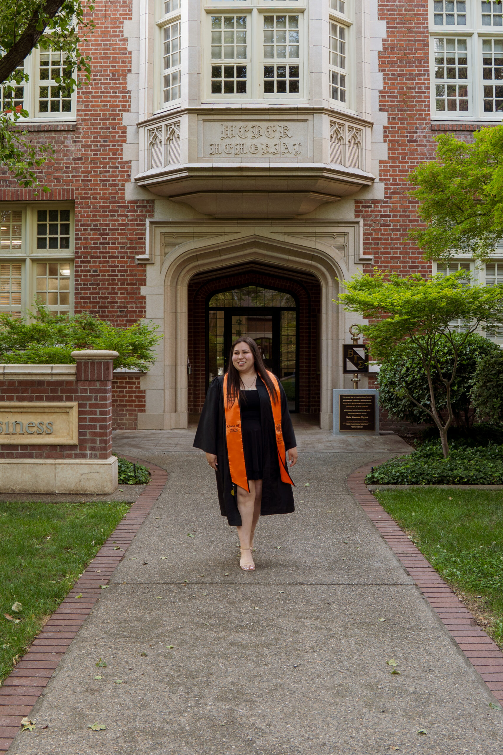 Alondra Duran in graduation attire walking on campus in front of a brick academic building